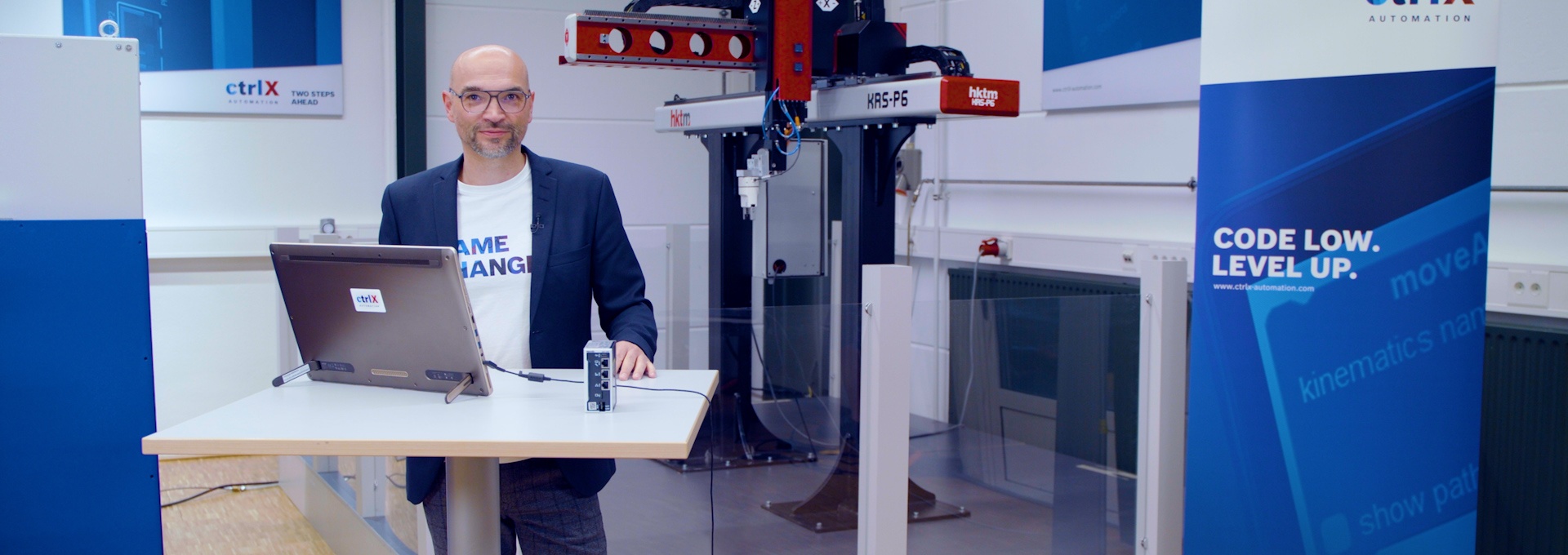 Gastgeber Christian Zentraf steht an einem Besprechungstisch und zeigt in einer Live-Demo, wie die Automatisierungslösung ctrlX AUTOMATION eine Pick-and-Place-Applikation steuert und antreibt
