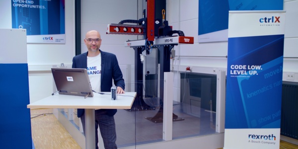 Gastgeber Christian Zentraf steht an einem Besprechungstisch und zeigt in einer Live-Demo, wie die Automatisierungslösung ctrlX AUTOMATION eine Pick-and-Place-Applikation steuert und antreibt
