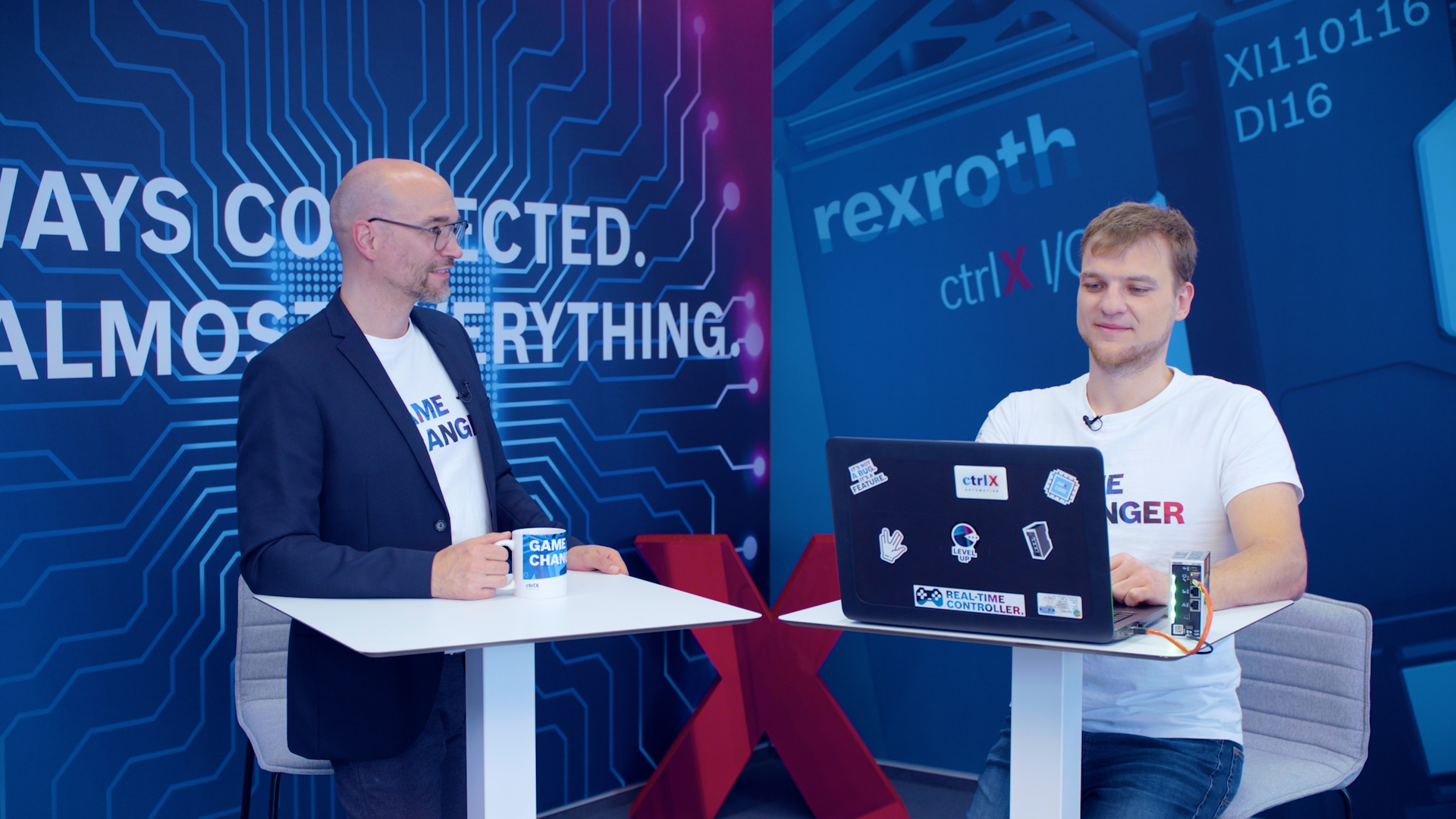 Gospodarz Christian Zentraf i jego gość Michael Schäfer stoją przy stołach i rozmawiają o programowaniu skryptów Pythona bezpośrednio na ctrlX CORE, sprzęcie sterującym rozwiązania automatyzacji ctrlX AUTOMATION.