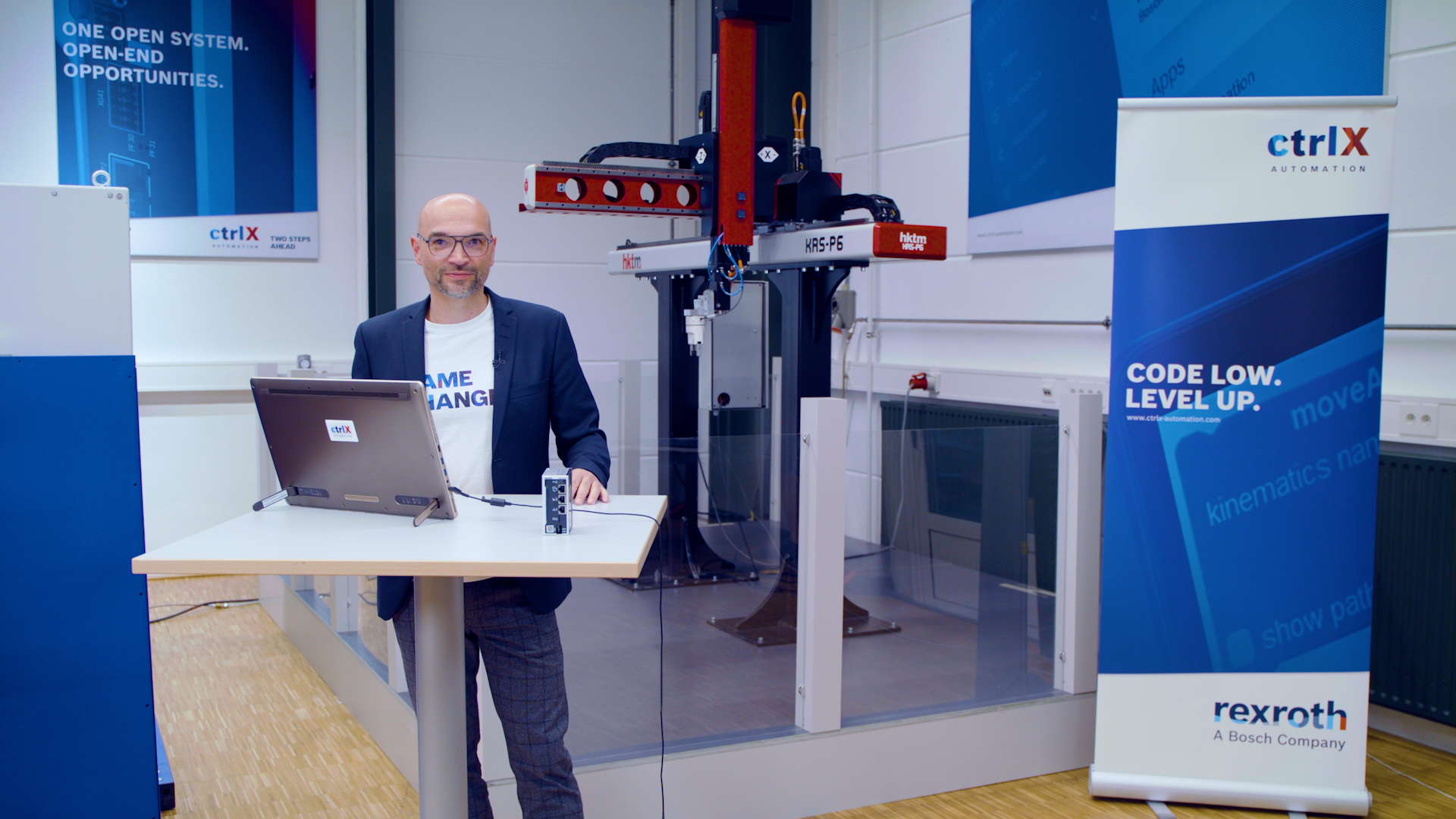 Gastgeber Christian Zentraf steht an einem Besprechungstisch und zeigt in einer Live-Demo, wie die Automatisierungslösung ctrlX AUTOMATION eine Pick-and-Place-Applikation steuert und antreibt